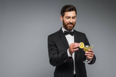 happy man in suit with bow tie holding sliced lemon and glass with cocktail isolated on grey