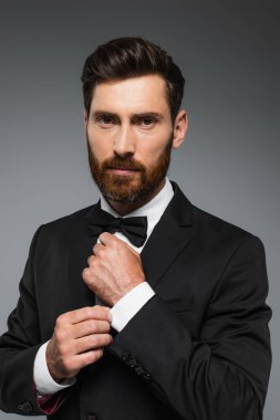 successful man in elegant suit adjusting bow tie isolated on grey 
