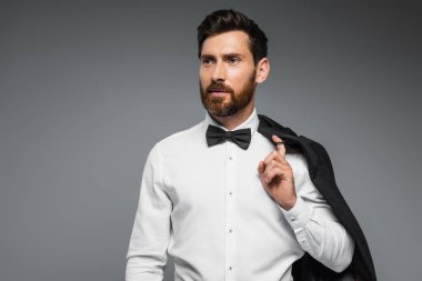 bearded man in white shirt with black bow tie holding blazer isolated on grey 