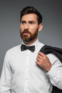 portrait of bearded man in suit with bow tie holding blazer isolated on grey 