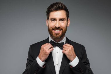 bearded man in suit smiling and adjusting bow tie isolated on grey 
