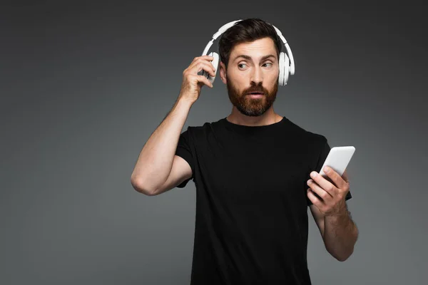 curious man adjusting wireless headphones and using smartphone isolated on grey