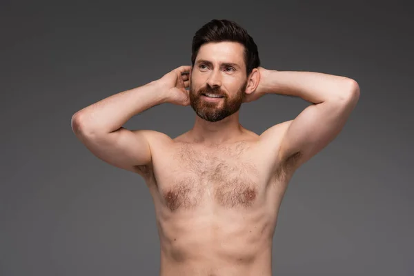 shirtless and happy man with hair on chest posing with hands behind head isolated on grey