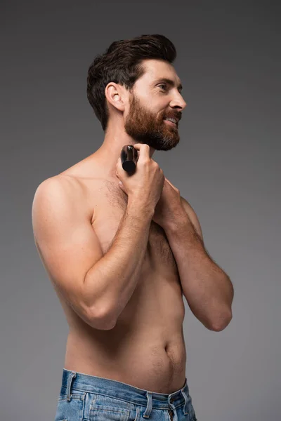 smiling and shirtless man shaving beard with electric razor isolated on grey