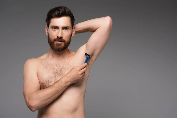 bearded and shirtless man shaving armpit with safety razor isolated on grey