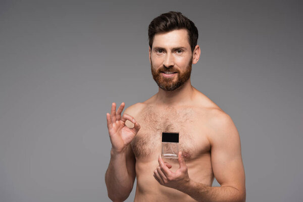 happy and shirtless man holding bottle with perfume and showing ok sign isolated on grey