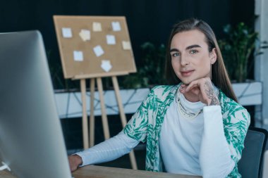 Tattooed queer designer looking at camera near computer in creative agency 