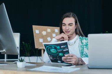 Positive queer designer looking at paper near gadgets on table in creative agency 