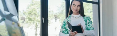 Smiling nonbinary person writing on notebook in creative agency, banner 
