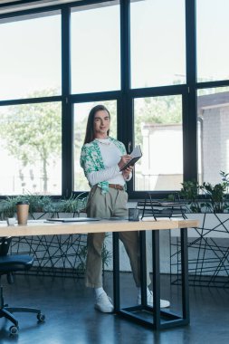Smiling queer person writing on notebook and looking at camera in creative agency 