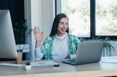 Cheerful queer designer having video call on laptop in creative agency 