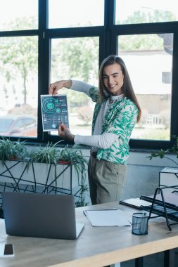 Cheerful queer designer holding paper with charts during video call on laptop in office 