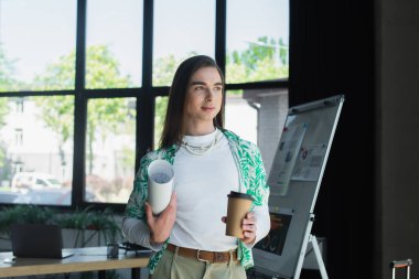 Brunette queer person holding blueprint and coffee to go in office 