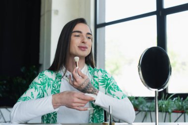Smiling queer person holding face powder near mirror in creative agency 