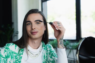 Queer designer applying mascara brush near blurred mirror in office 