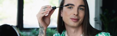 Nonbinary designer holding mascara brush and looking at camera in office, banner 