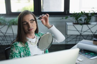 Nonbinary designer applying mascara near mirror in creative agency 