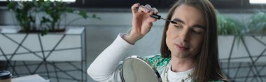 Nonbinary designer applying mascara near mirror in creative agency, banner 