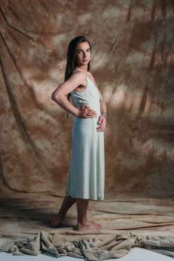 Full length of barefoot queer person in sundress looking at camera on abstract brown background 