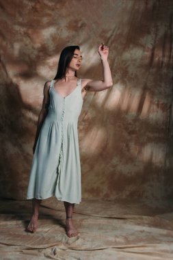 Barefoot nonbinary person in light blue sundress posing on abstract brown background 