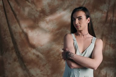 Young queer person in sundress looking at camera on abstract brown background 