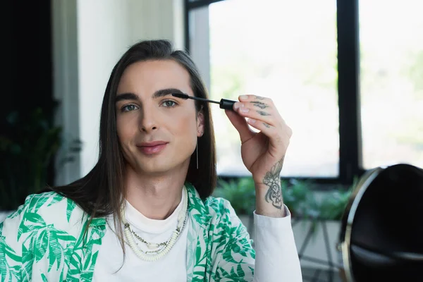 Queer designer applying mascara brush near blurred mirror in office