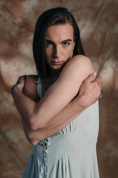 Portrait of nonbinary person in sundress touching shoulders on abstract brown background