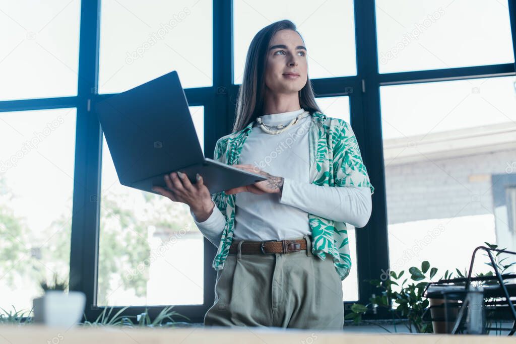 Low angle view of nonbinary person holding laptop in creative agency 