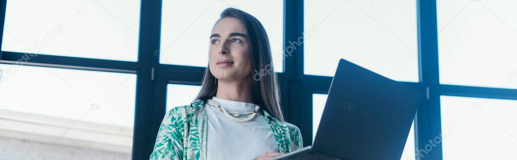 Low angle view of queer designer holding laptop in office, banner 
