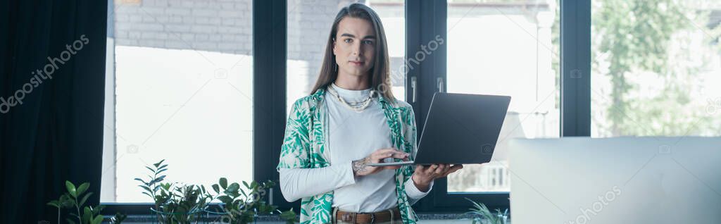 Queer designer holding laptop and looking at camera in creative agency, banner 