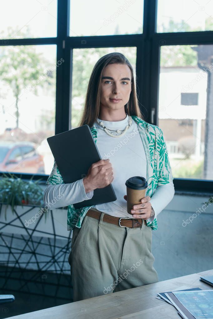 Queer person holding laptop and coffee to go and looking at camera in creative agency 