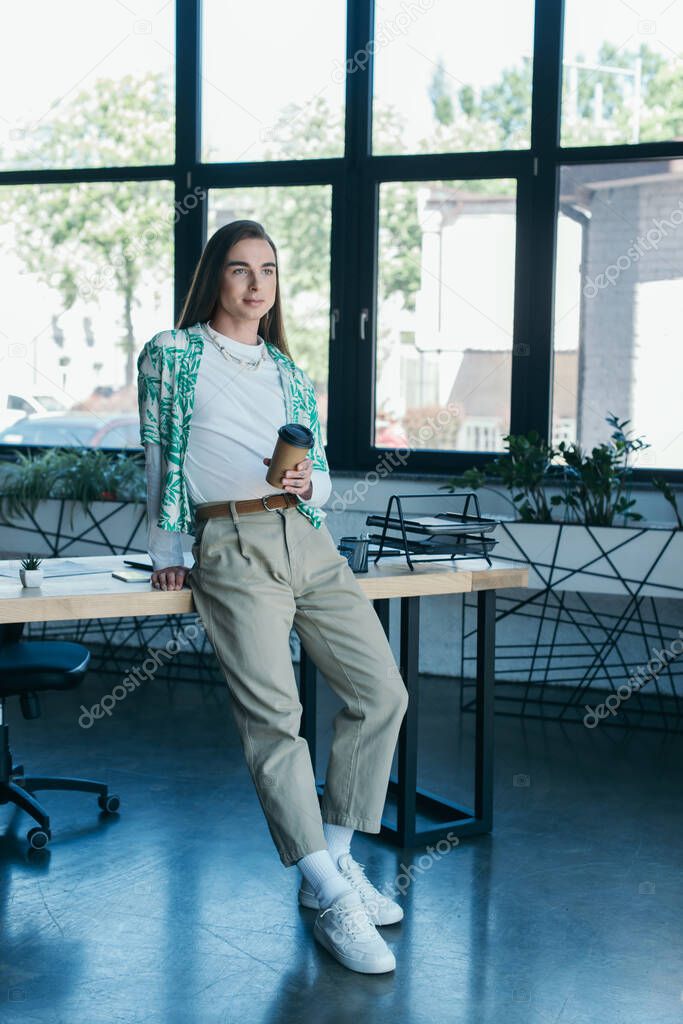 Young queer designer holding coffee to go near working table in office 
