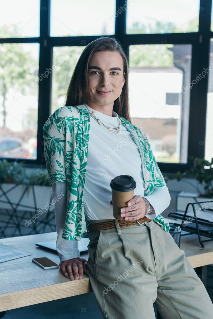 Smiling queer designer holding paper cup near working table in creative agency 
