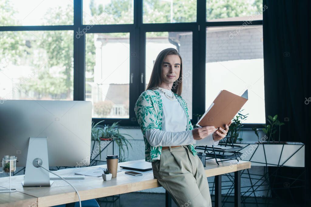 Smiling queer designer holding paper folder and looking at camera near working table in office 