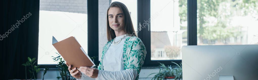 Young queer designer holding paper folder and looking at camera near computer in office, banner 