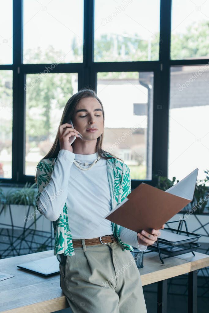 Queer designer holding paper folder and talking on smartphone in office 