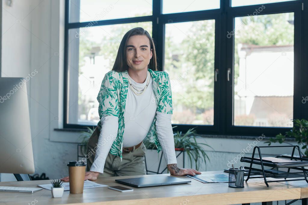 Positive nonbinary designer looking at camera near devices and coffee in creative agency 