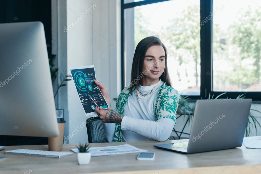 Smiling nonbinary designer holding charts during video call on laptop in office 
