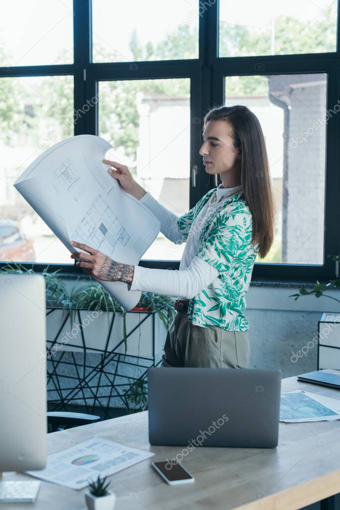 Side view of queer designer holding blueprint near devices in office 