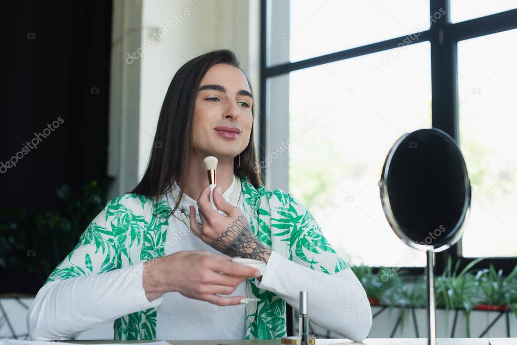 Smiling queer person holding face powder near mirror in creative agency 
