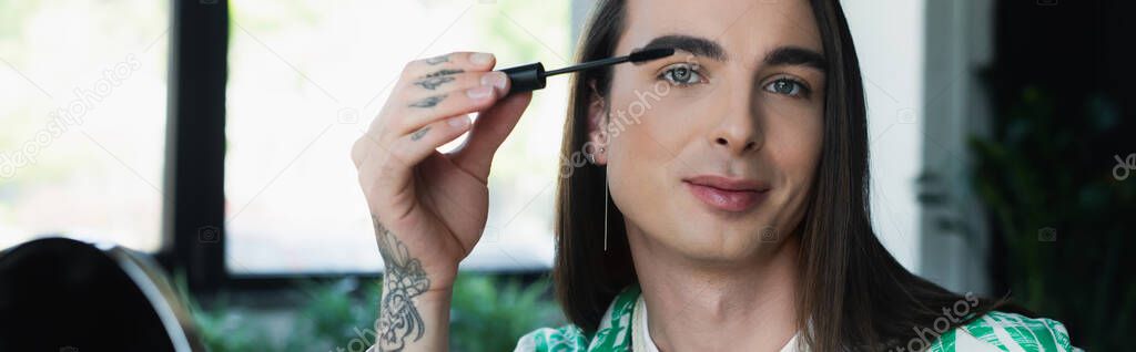 Nonbinary designer holding mascara brush and looking at camera in office, banner 