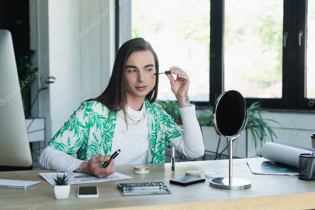 Queer designer holding mascara near decorative cosmetics and devices in office 