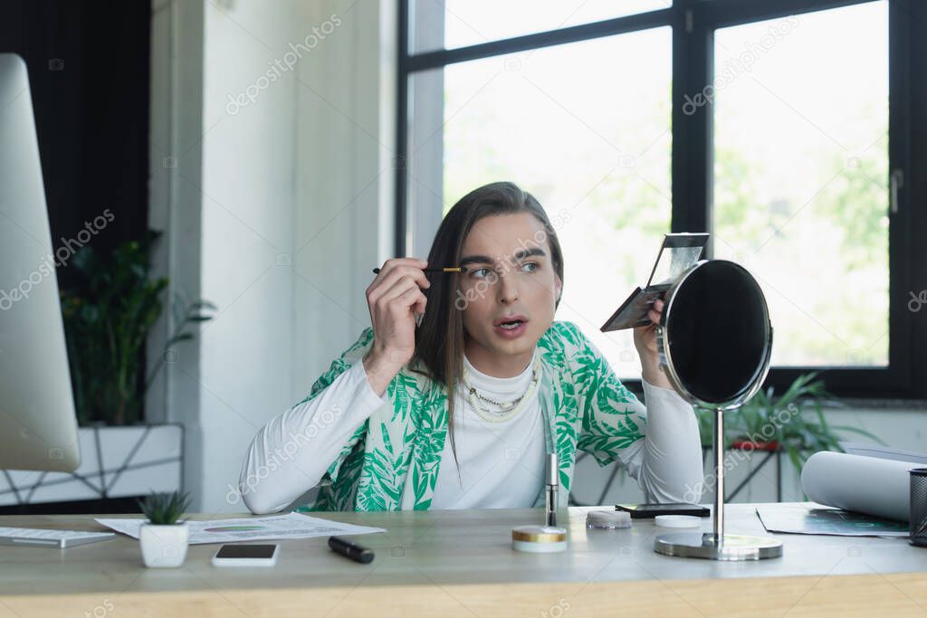 Queer person applying eye shadow near mirror and gadgets in office 