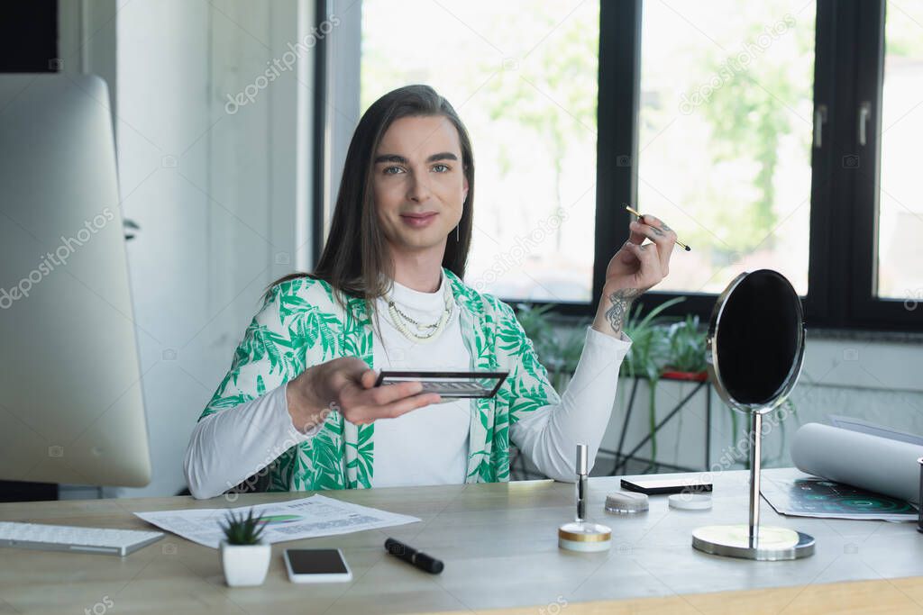 Smiling queer designer holding eye shadow near gadgets and mirror in office 