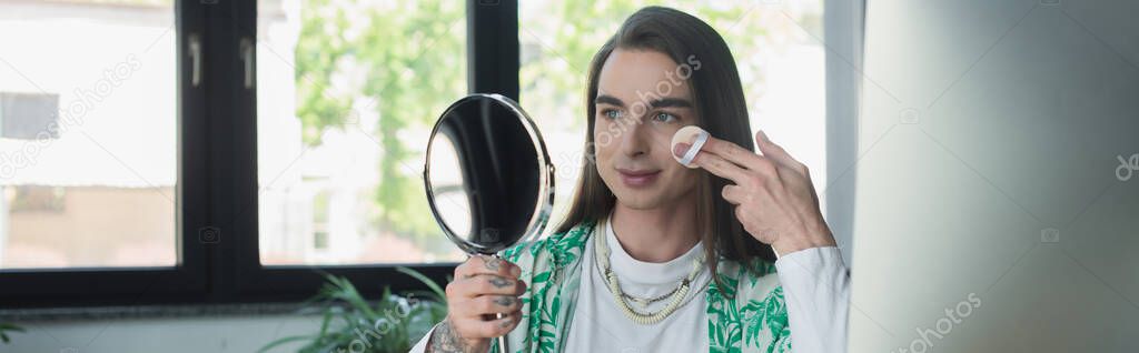 Queer designer applying face powder and holding mirror in office, banner 