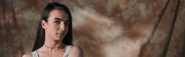 Brunette queer person looking at camera on abstract brown background, banner 