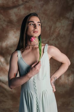 Brunette nonbinary person in sundress holding tulip on abstract brown background 