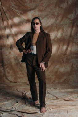 Full length of stylish queer person in suit and corset standing on abstract brown background