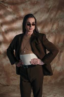 Brunette queer person in sunglasses holding hands on hips on abstract brown background clipart