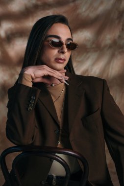 Trendy queer person in sunglasses and blazer sitting on chair on abstract brown background